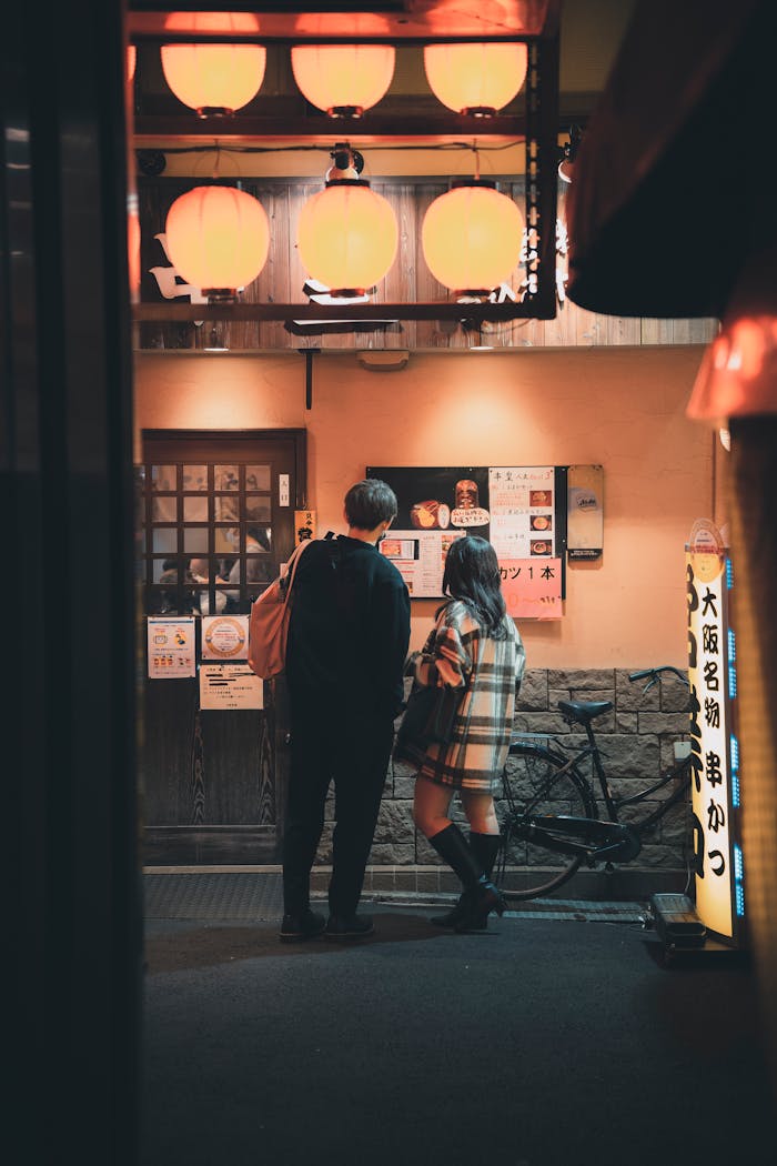 Couple inspecting menu outside cozy, lamp-lit Japanese restaurant at night.